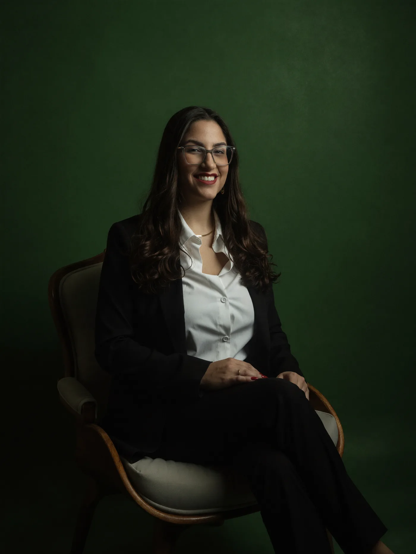 Jennifer Paim sorrindo, sentada em uma poltrona clássica diante de um fundo verde